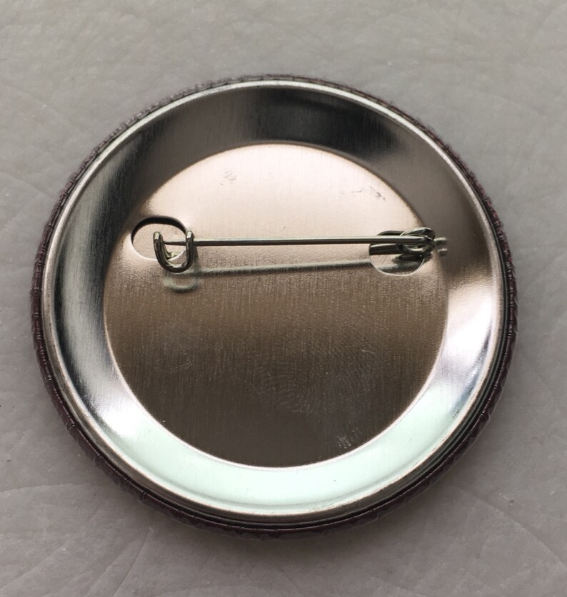 May include: The back of a round button with a silver metal backing and a pin attached.
