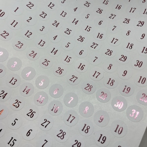 May include: A sheet of white circular stickers featuring printed numbers from 1 to 31 in a metallic rose gold foil finish. The numbers are arranged in multiple repeating rows and columns across the adhesive paper, providing a set of calendar dates.