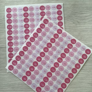 May include: Two sheets of pink and light pink circular stickers with black text indicating weight loss in pounds, ranging from -0.23kg to -2.27kg. The stickers are on a light wood-grain surface.