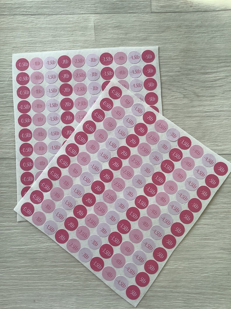 May include: Two sheets of pink and light pink circular stickers with black text indicating weight loss in pounds, ranging from -0.23kg to -2.27kg. The stickers are on a light wood-grain surface.