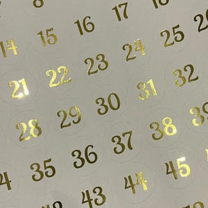 May include: A sheet of circular adhesive stickers featuring sequential numbers printed in a metallic gold foil finish. The numbers are arranged in a grid pattern on a plain white background, with each digit centred within a faint circular outline.