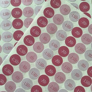 May include: Circular stickers in varying shades of pink and red, each printed with a weight measurement in pounds. The stickers range from -0.5lb to -5lb. The stickers are arranged on a light-colored sheet.