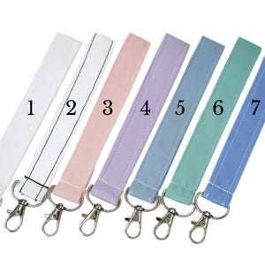 May include: Seven fabric wristlet keychains arranged in a row, numbered one through seven. The colors include white, white with black stripes, light pink, lavender, light blue, mint green, and medium blue, each featuring a silver metal lobster clasp.