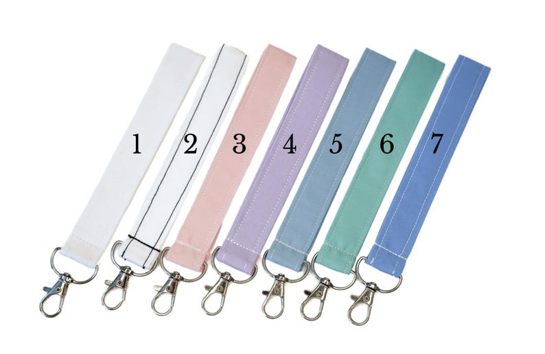 May include: Seven fabric wristlet keychains arranged in a row, numbered one through seven. The colors include white, white with black stripes, light pink, lavender, light blue, mint green, and medium blue, each featuring a silver metal lobster clasp.