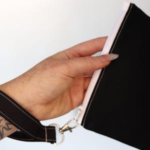 May include: A black fabric wristlet pouch featuring a white zipper closure and a matching black wrist strap attached with a silver-toned metal swivel clasp. The pouch is constructed from solid black material with visible white stitching details.