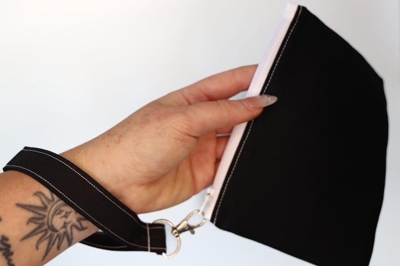 May include: A black fabric wristlet pouch featuring a white zipper closure and a matching black wrist strap attached with a silver-toned metal swivel clasp. The pouch is constructed from solid black material with visible white stitching details.