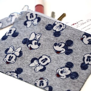 May include: A gray fabric zipper pouch featuring a repeating pattern of dark blue Mickey and Minnie Mouse character faces. The pouch includes a matching gray fabric wrist strap attached to the side and contains a business card and pens.
