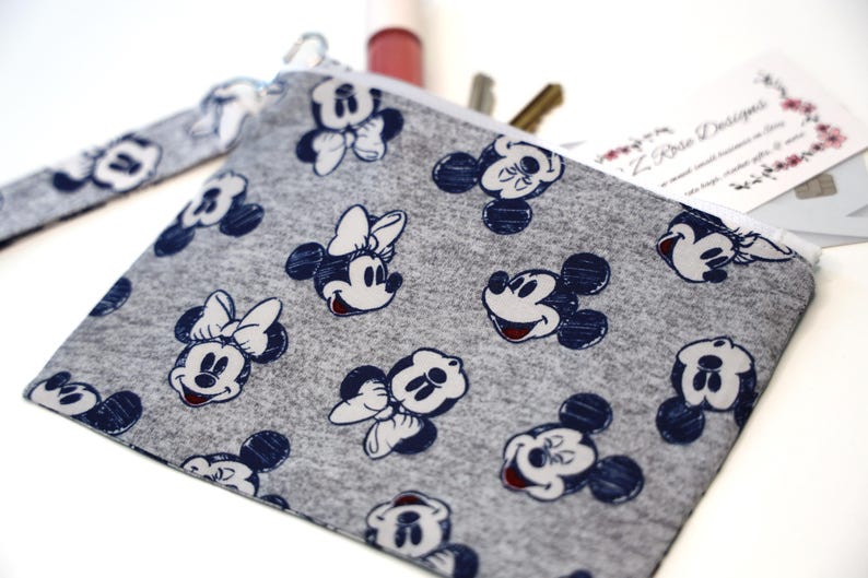 May include: A gray fabric zipper pouch featuring a repeating pattern of dark blue Mickey and Minnie Mouse character faces. The pouch includes a matching gray fabric wrist strap attached to the side and contains a business card and pens.