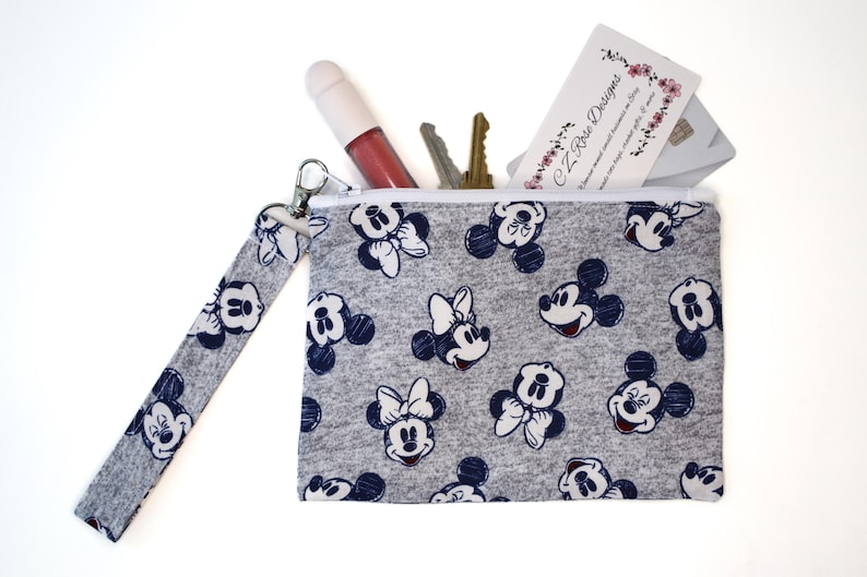 May include: A gray zippered wristlet pouch featuring a repeating pattern of Mickey and Minnie Mouse faces in dark blue. The pouch includes a matching wrist strap and contains a lip gloss, two metal keys, and a business card labeled CZ Rose Designs.