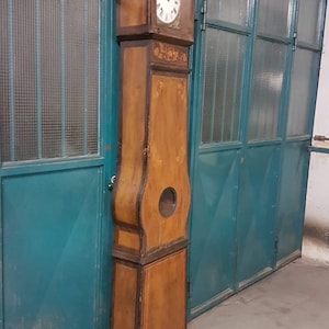 Puede incluir: Un reloj de abuelo de madera antiguo con un acabado marrón claro. El reloj presenta una esfera blanca, tallas decorativas y una abertura circular en el cuerpo. El reloj está colocado contra una pared de color verde azulado.