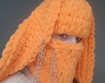 May include: A bright orange crocheted mask with long ear flaps and silver chains attached. The mask covers the face and has a fluffy texture.