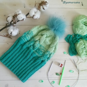 May include: A green and light green knitted hat with a fluffy blue pom pom. The hat is on a white wooden surface with cotton branches, yarn, and knitting needles.