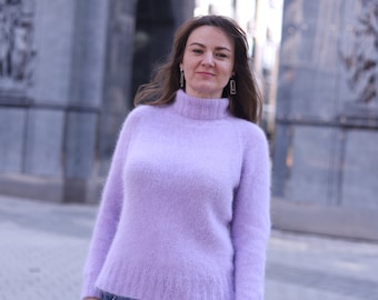 May include: A woman wearing a light purple fuzzy sweater and blue jeans stands in front of a stone archway.