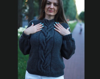 May include: A dark gray cable knit sweater with a high neckline. The sweater has a braided design down the front.