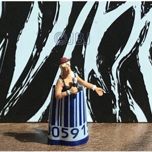 May include: A small figurine of a woman wearing a black top, a red hat, and holding a microphone. She is standing inside a blue and white striped tube with the number "0591" printed on it.