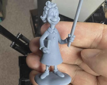 May include: A gray figurine of a cartoon character, holding a book and a sign. The character is wearing a dress and has curly hair. The figurine is standing on a round base.