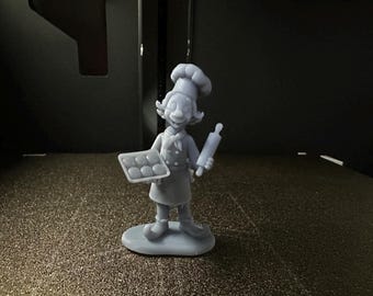 May include: A light gray 3D printed figurine of a chef wearing a tall toque and apron. The character holds a rectangular baking tray with six circular items in one hand and a rolling pin in the other, standing on a smooth oval base.