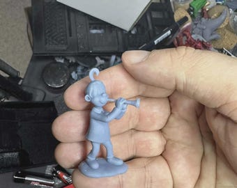 May include: A light blue figurine of a person playing a trumpet. The figurine is standing on a round base. The person is wearing a short-sleeved shirt and pants. The figurine is held in a hand.