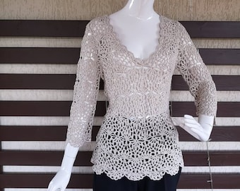 May include: A beige crocheted top with a v-neck and long sleeves. The top has a scalloped hem and a delicate crocheted pattern.