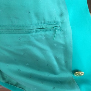 May include: A teal green blazer with a zippered pocket and a gold button. The lining of the blazer is teal green with a repeating pattern of the letter "R".