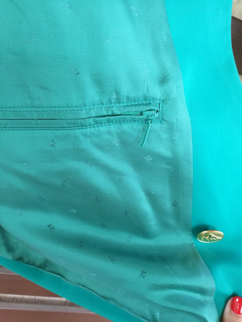 May include: A teal green blazer with a zippered pocket and a gold button. The lining of the blazer is teal green with a repeating pattern of the letter "R".
