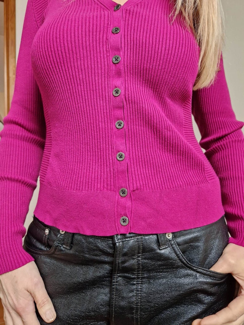 May include: A close-up of a woman wearing a pink ribbed cardigan with a button-up front and black leather pants.