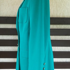 May include: A teal blazer with gold buttons on the sleeve. The blazer is on a mannequin and is photographed against a wood and white background.