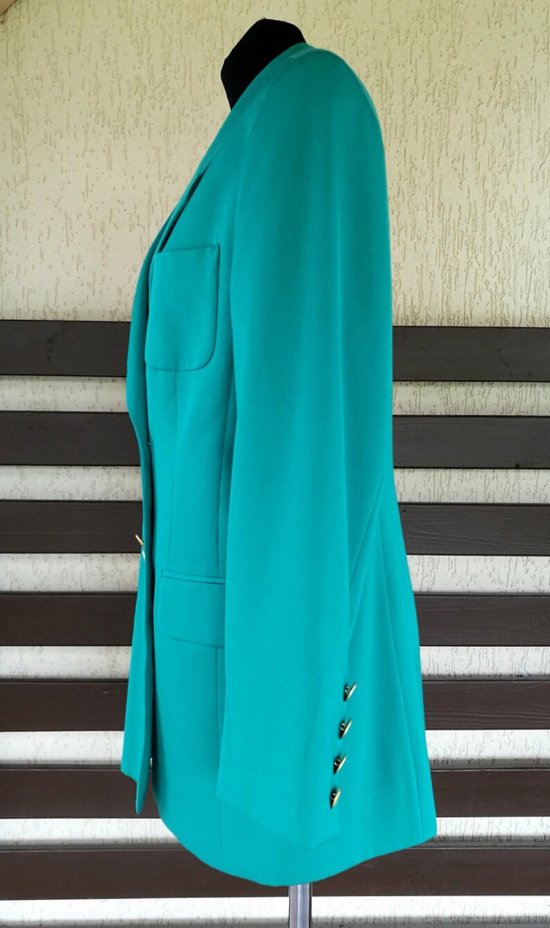 May include: A teal blazer with gold buttons on the sleeve. The blazer is on a mannequin and is photographed against a wood and white background.