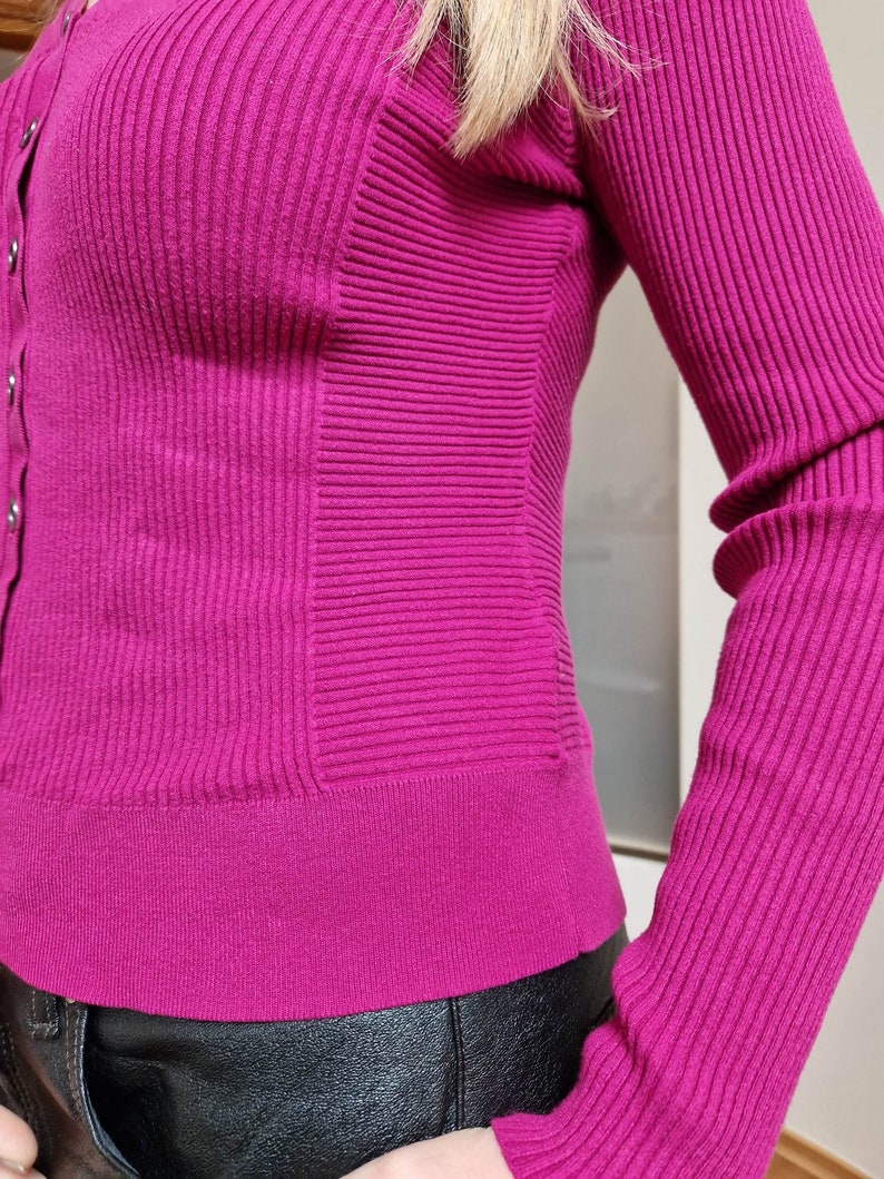 May include: A close-up of a magenta ribbed cardigan with a button-up front and long sleeves. The cardigan is made of a soft, stretchy material and has a fitted silhouette.