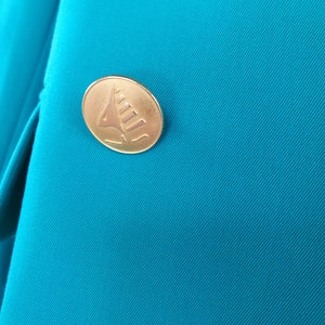 May include: A close-up of a teal blue fabric with a gold button featuring a stylized sailboat design.