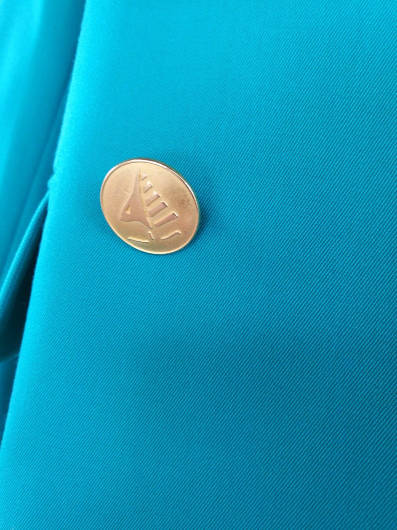 May include: A close-up of a teal blue fabric with a gold button featuring a stylized sailboat design.