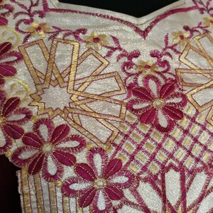 May include: A close-up of a white fabric with gold and burgundy embroidery. The embroidery features a geometric pattern with flowers and stars.
