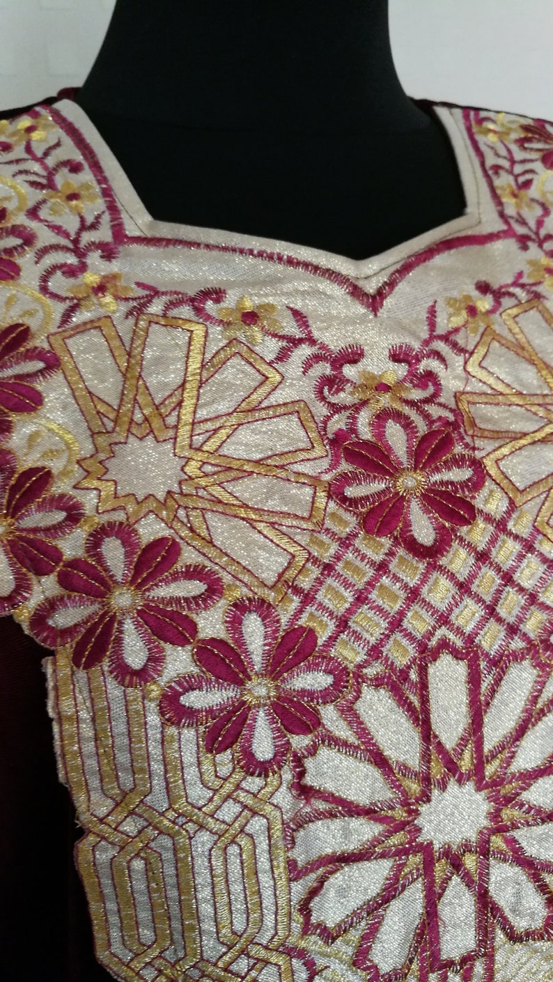 May include: A close-up of a white fabric with gold and burgundy embroidery. The embroidery features a geometric pattern with flowers and stars.