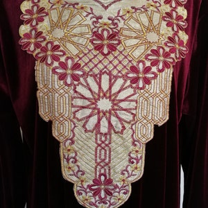 May include: A burgundy velvet dress with gold and burgundy embroidery around the neckline. The embroidery features a geometric pattern with floral accents.