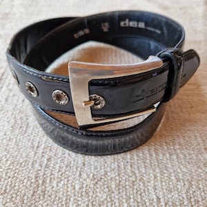 Pode incluir: Um cinto de couro preto com uma fivela de prata. A fivela tem a palavra "dea" gravada. O cinto tem um acabamento brilhante e é decorado com ilhós de prata.