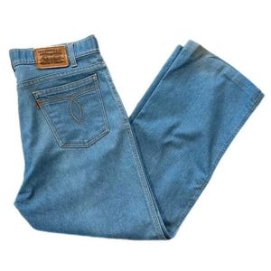May include: Light blue denim jeans with a classic five-pocket design. The jeans feature a straight leg cut and a small red tag on the back pocket. A brown leather patch with the Levi Strauss & Co. logo is visible on the waistband.