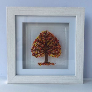 May include: A framed artwork featuring a tree with a brown trunk and a full crown of autumn-colored leaves in shades of red, orange, and yellow. The artwork is set against a light background and framed in a white wooden frame.