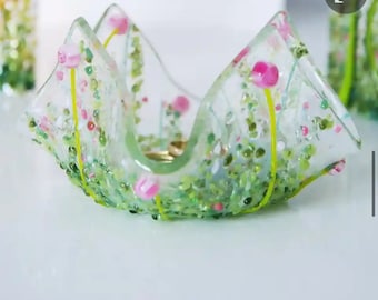 May include: A clear glass bowl with a floral design. The bowl has a wavy shape and is decorated with pink flowers, green leaves, and small green and pink accents. The stems are yellow. The bowl is sitting on a white surface.