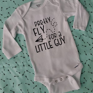 May include: White baby onesie with long sleeves. The text "Pretty Fly for a Little Guy" is printed in black, along with a paper airplane graphic and dotted lines. The onesie has snap closures at the bottom.