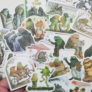 May include: A collection of colorful Frog and Toad stickers featuring illustrations of the characters in various scenes. The stickers include images of the characters reading, riding a bicycle, and more. The text "Frog and Toad" is visible.