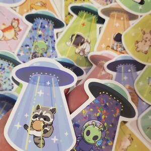May include: A set of colorful stickers featuring cartoon animals and aliens being abducted by UFOs. The stickers are printed on white paper and have a glossy finish.