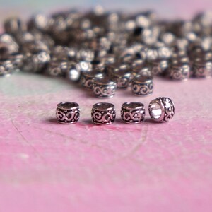 May include: Five silver metal beads with a decorative swirl design. The beads are arranged in a row on a pink surface.