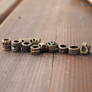 May include: A collection of ten metal beads with various designs, including a skull, a peace sign, and a swirl pattern. The beads are a bronze color and are arranged in a row on a wooden surface.