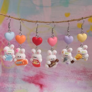 May include: Six pairs of dangle earrings. Each pair features a white bunny figure with different accessories, suspended below a heart-shaped charm. The charms are in blue, orange, red, pink, purple, and yellow. The earrings hang from a brown string.