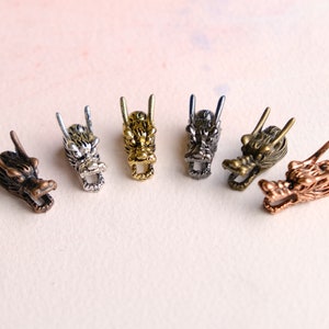 May include: Five dragon head charms in different finishes: silver, gold, bronze, and two shades of antique brass. The dragon heads are detailed with scales and have open mouths.