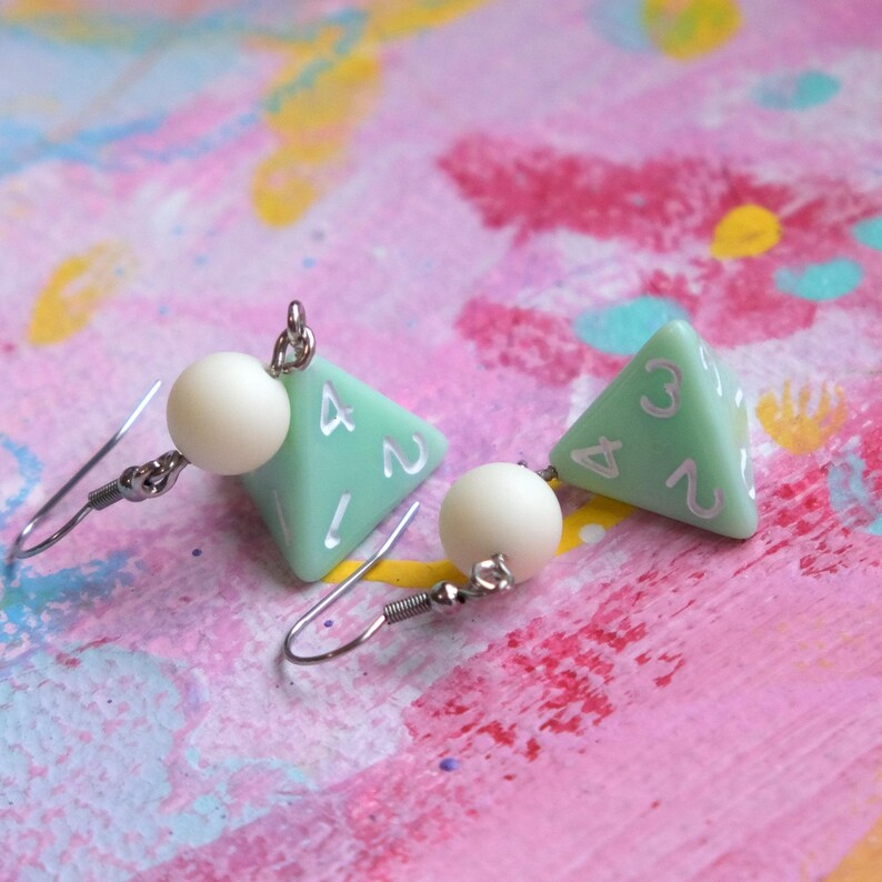 May include: A pair of silver dangle earrings with a green four-sided dice charm and a white pearl bead on each earring.