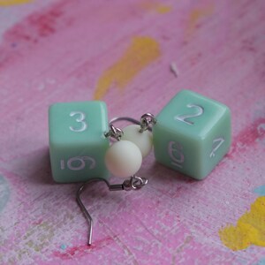 May include: A pair of earrings featuring two light green dice with white numbers. The dice are connected by a silver chain and a white bead.