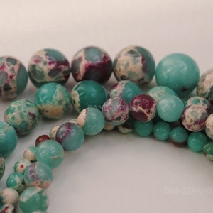 May include: A strand of green and white jasper beads with a mix of round and oval shapes. The beads have a mottled pattern with hints of purple.