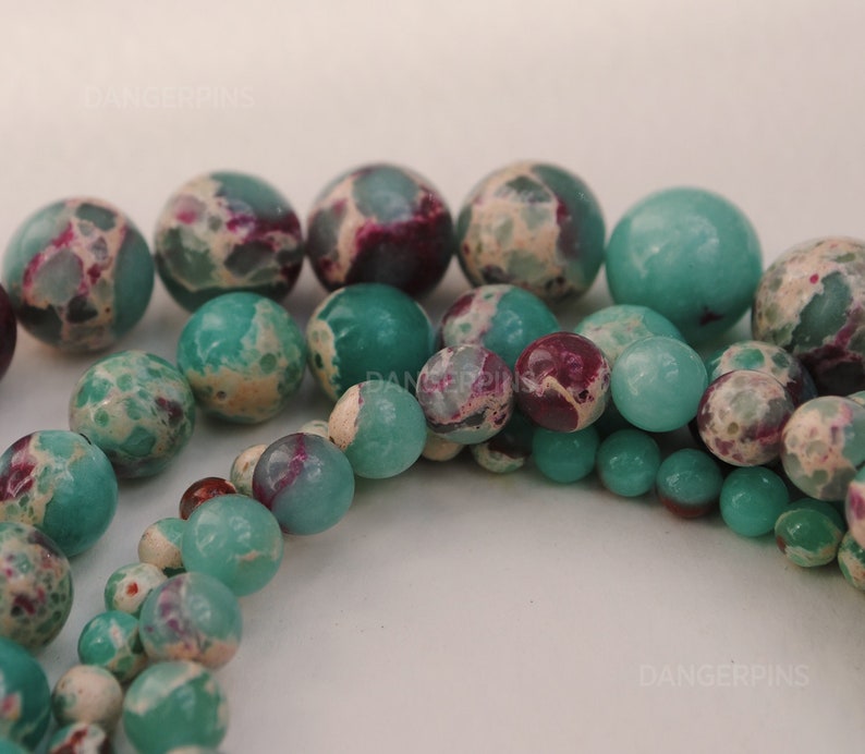 May include: A strand of green and white jasper beads with a mix of round and oval shapes. The beads have a mottled pattern with hints of purple.