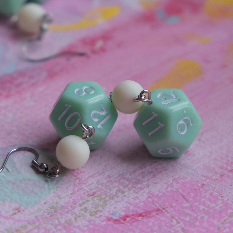 May include: A pair of green dice earrings with white numbers. Each earring features a white pearl bead and a silver hook.